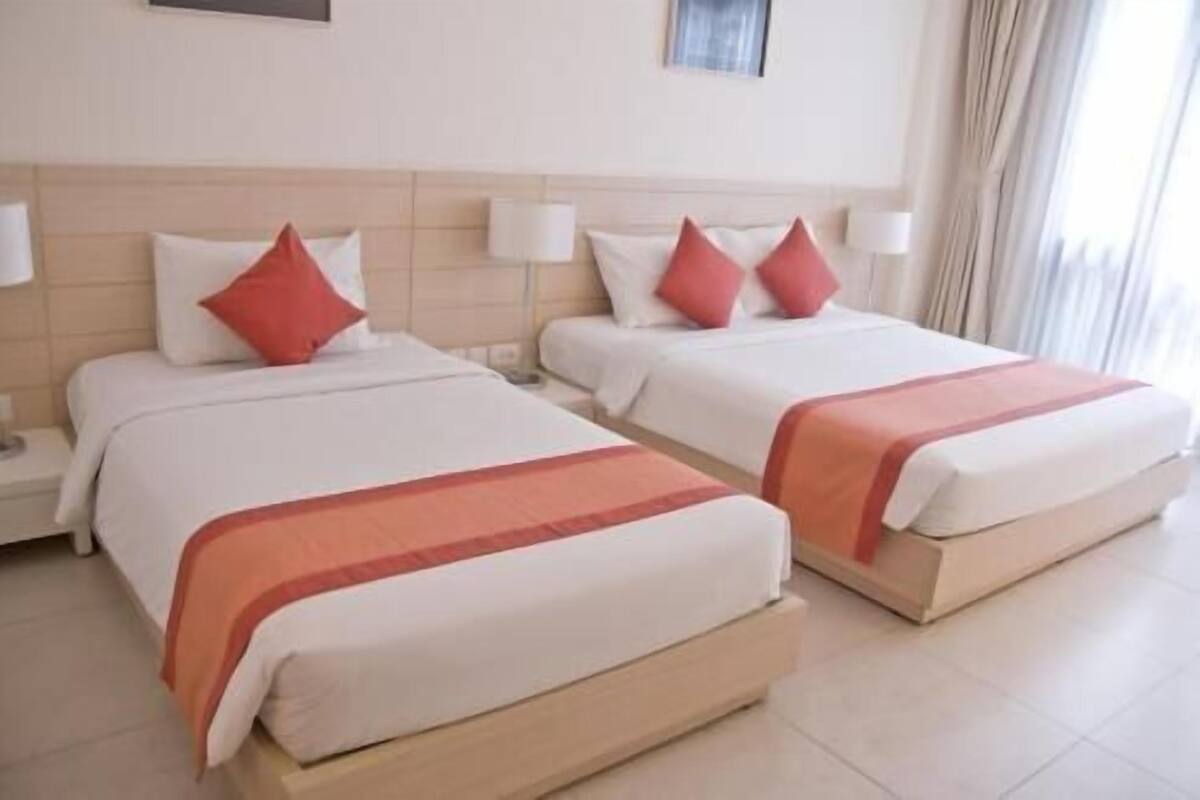 Dos camas individuales con almohadas rojas y ropa de cama blanca, una mesita de noche con lámpara y una ventana con cortinas.