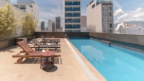 Un área de piscina en la azotea con sillones, una mesa con una bebida y una vista panorámica de la ciudad al fondo.