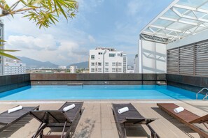 Una piscina en la azotea con sillones de descanso y vistas a un paisaje urbano y montañas.