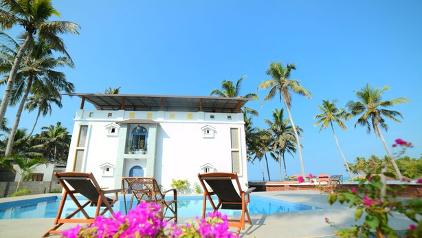 A white building with a pool, surrounded by palm trees and featuring two lounge chairs. - Magnolia Guest House (Varkala)