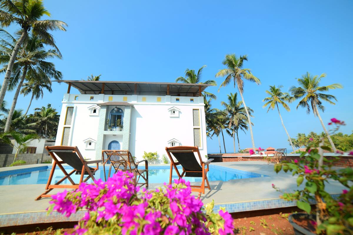 A white building with a pool, surrounded by palm trees and featuring two lounge chairs.