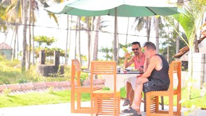A wooden table with chairs and a green umbrella, set outdoors with palm trees in the background. - Magnolia Guest House (Varkala)