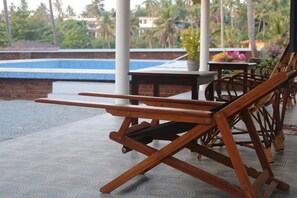 Outdoor wooden chairs and tables on a patio with a pool and palm trees in the background. - Magnolia Guest House (Varkala)