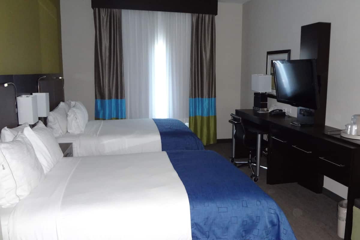 A hotel room with two beds, a desk with a computer, a TV, and a large window with curtains.
