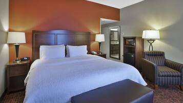 A hotel room with a large bed, two bedside tables with lamps, a chair, a bench, and a small desk with a lamp.