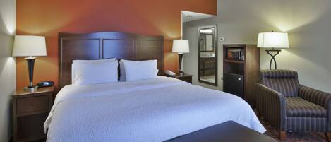 A hotel room with a large bed, two bedside tables with lamps, a chair, a bench, and a small desk with a lamp.