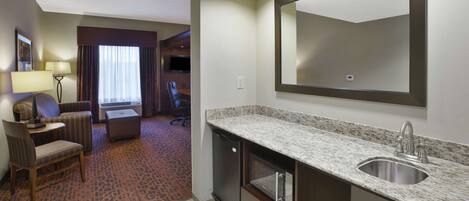 A hotel room with a kitchenette, a large mirror, a seating area with a chair and a small table, and a television in the background.