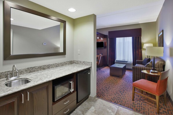 A hotel room with a kitchen area, a seating area with a sofa and armchair, and a large mirror.