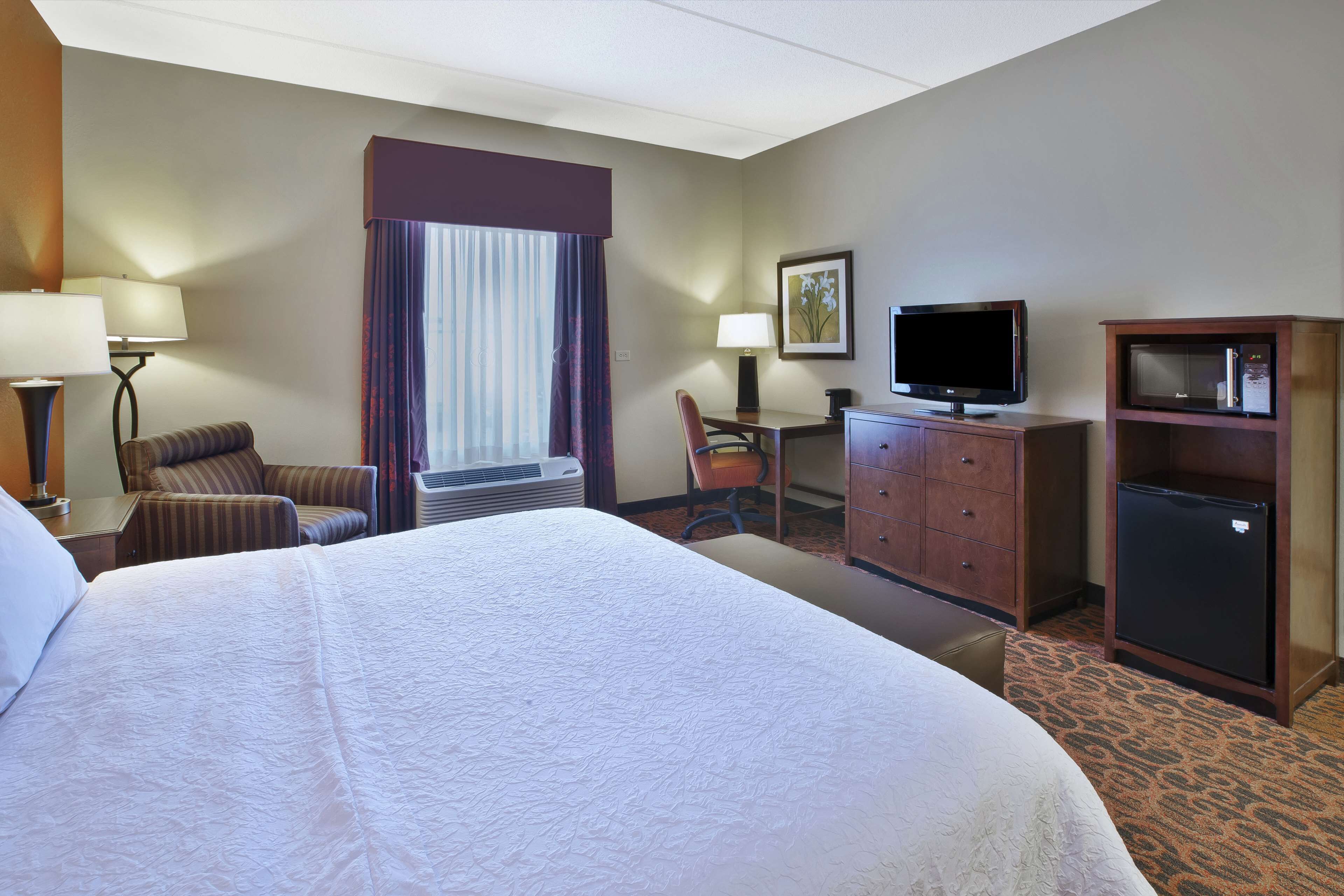 Ein Hotelzimmer mit einem großen Bett, einem Schreibtisch mit Stuhl, einem Fernseher, einem Kühlschrank und einem Fenster mit Vorhängen.