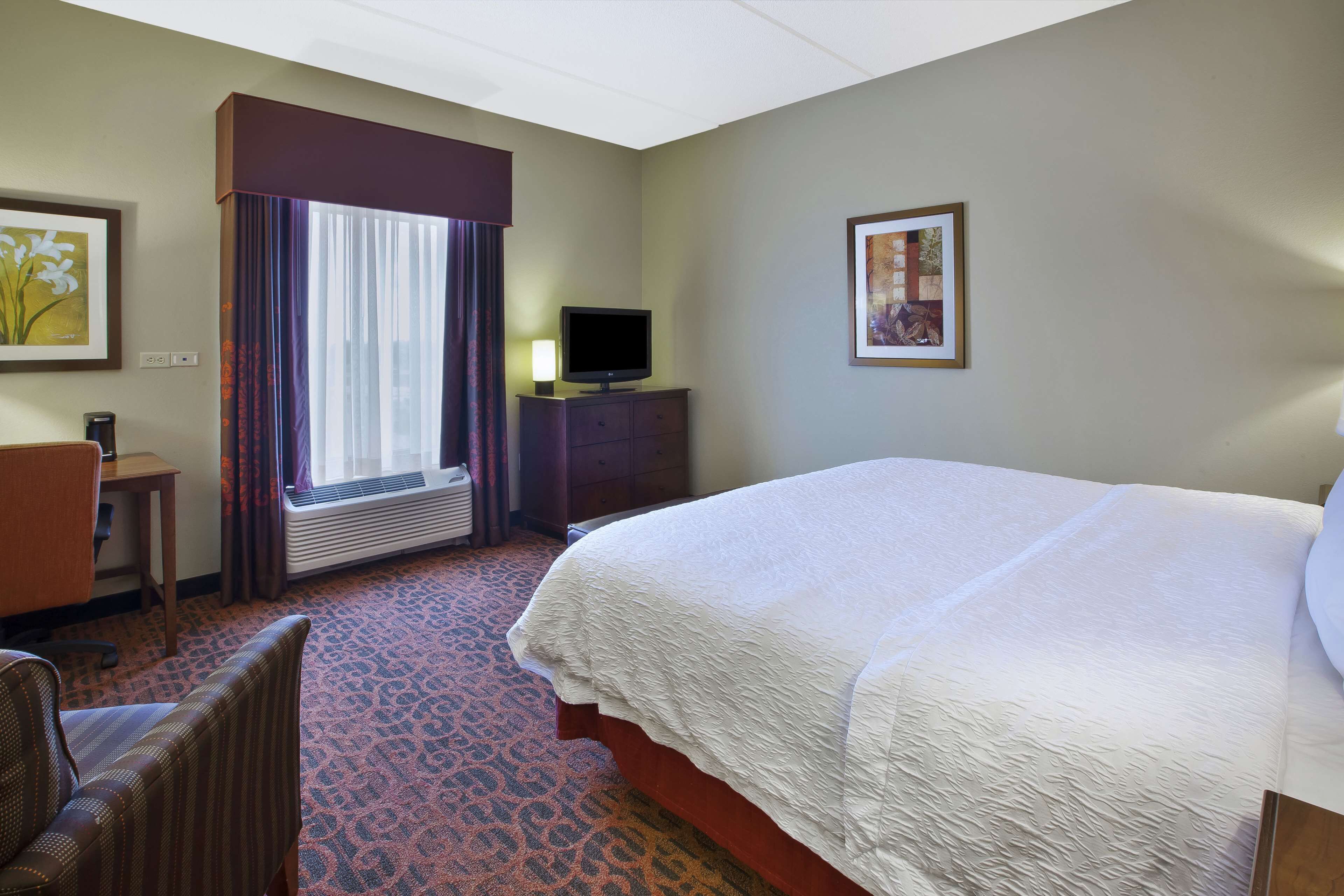 A hotel room with a large bed, a desk with a chair, a television on a dresser, and two framed pictures on the walls.