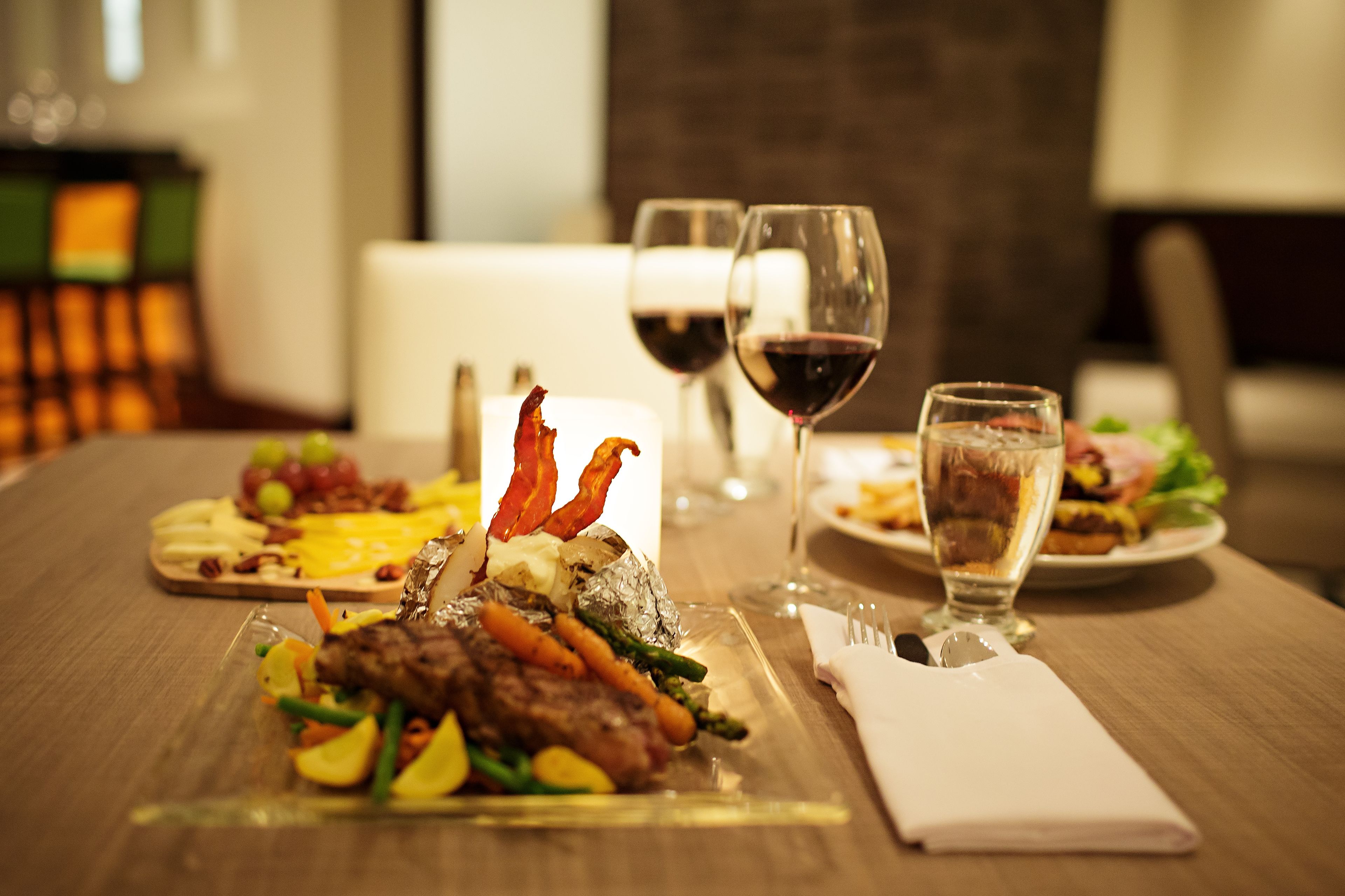 A dining table with a variety of dishes, including steak, vegetables, and a dessert. There are also two glasses of red wine and a water glass.