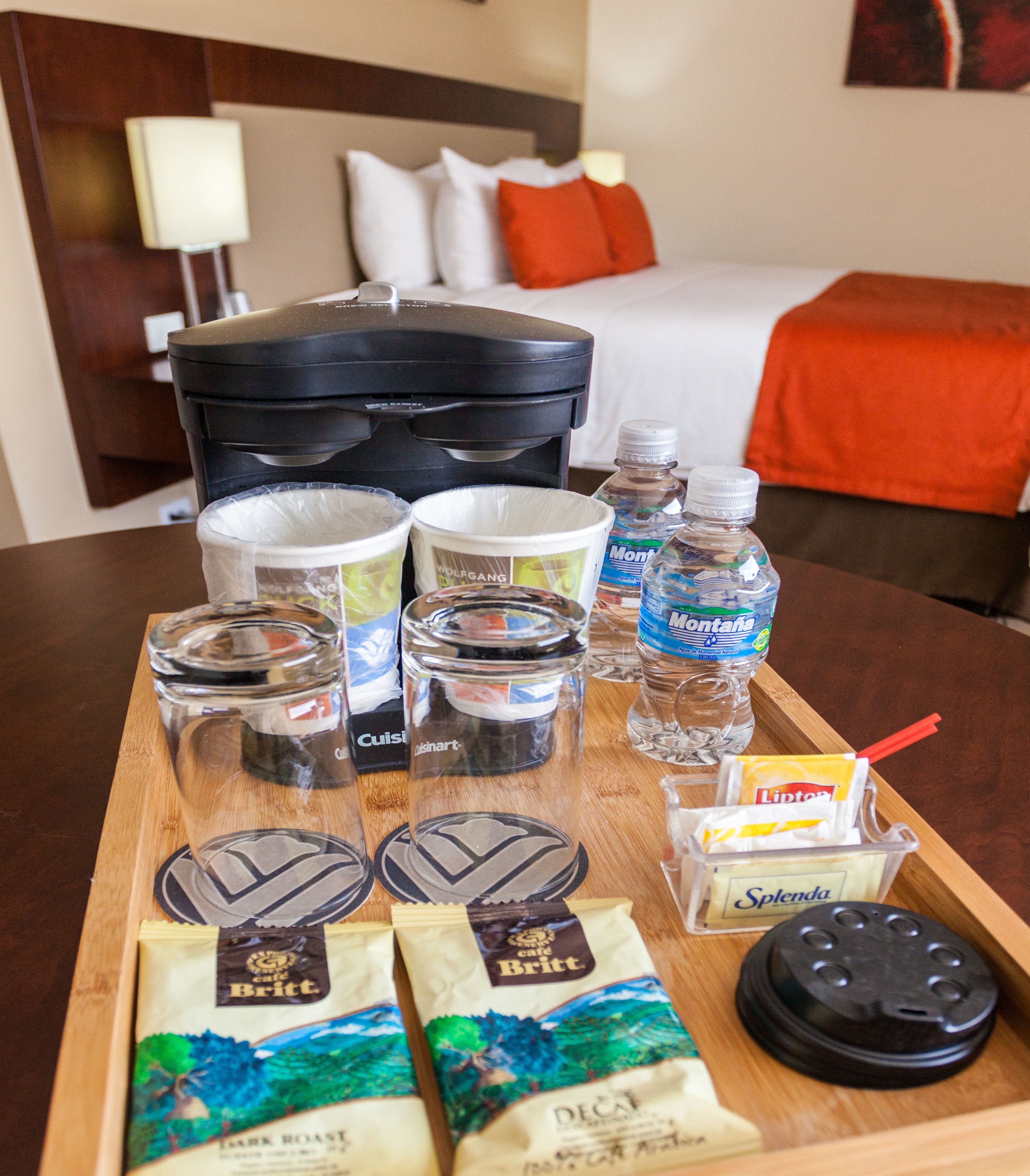 A hotel room with a bed, a bedside table with a coffee maker, water bottles, and coffee packets.