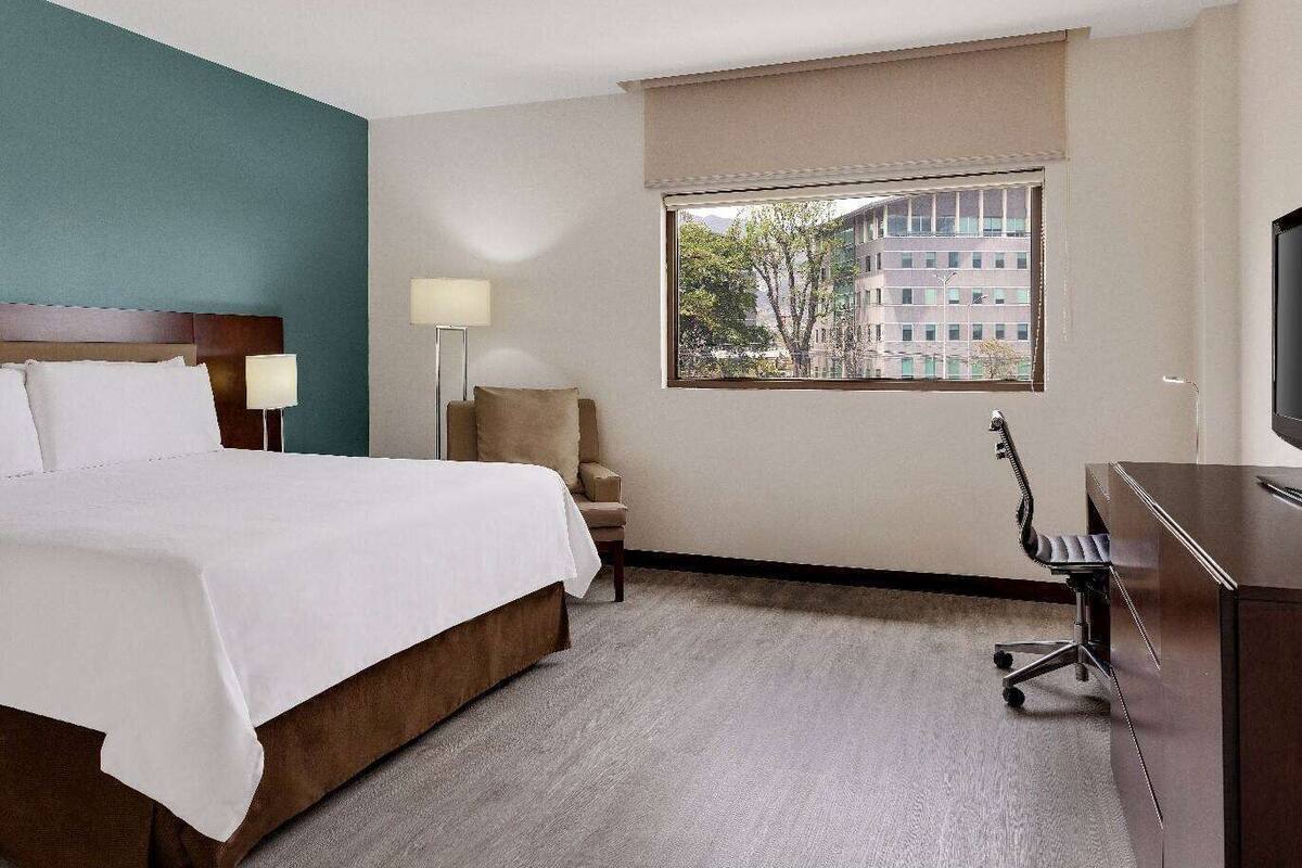 Hotelkamer met een bed, een bureau, een stoel, een lamp en een raam met uitzicht op gebouwen.