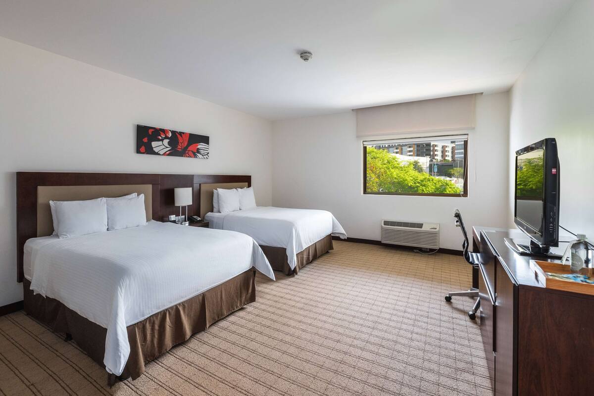 Een hotelkamer met twee bedden, een bureau met een computer, een televisie en een raam met uitzicht op gebouwen en bomen.