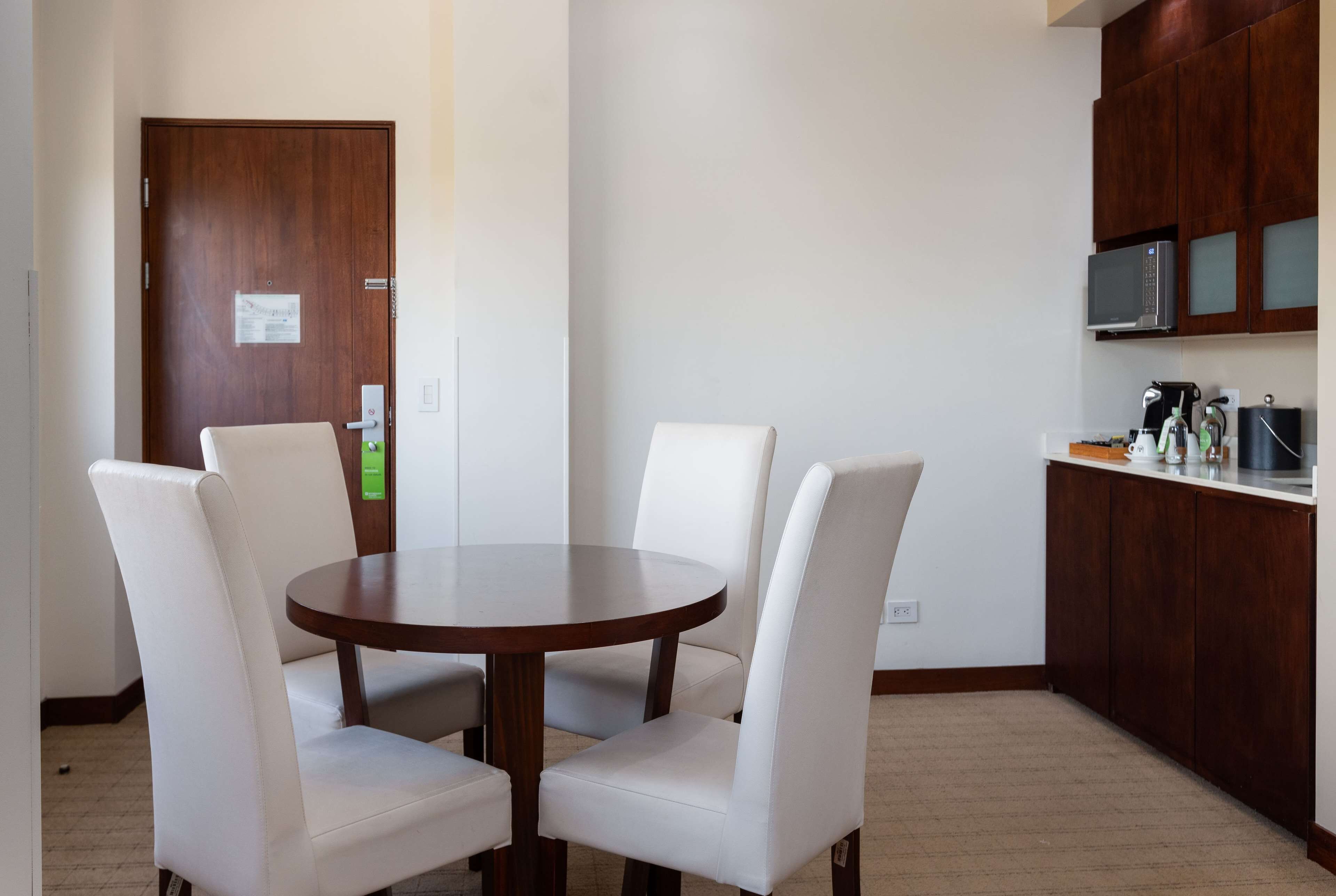 A dining area with a round table and four white chairs, a wooden door, and a kitchen with dark wood cabinets and a microwave.