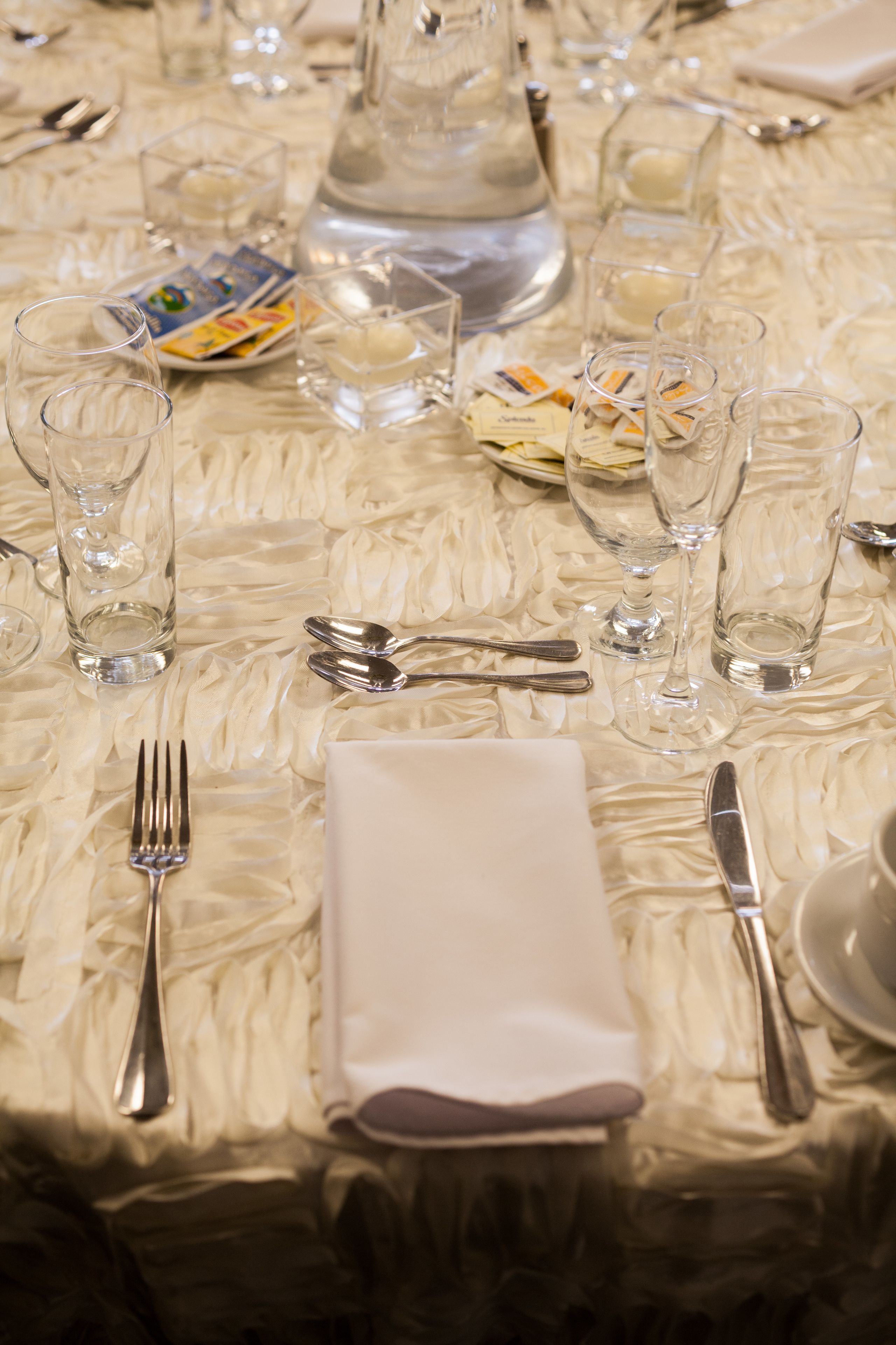 A table set with clear glassware, silverware, and folded napkins on a textured tablecloth.