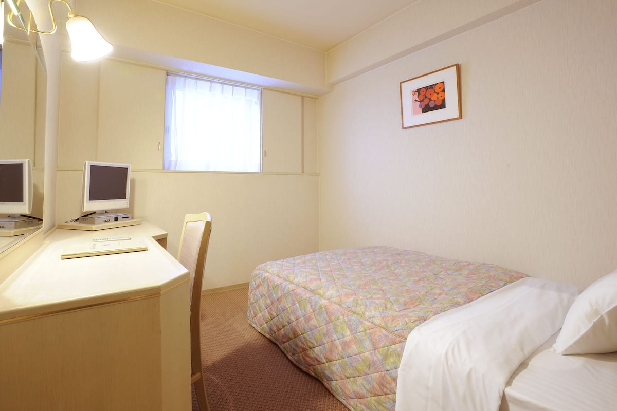 Hotellihuone, jossa on sänky, työpöytä tietokoneella, tuoli, ikkuna ja seinällä oleva kehystetty kuva.