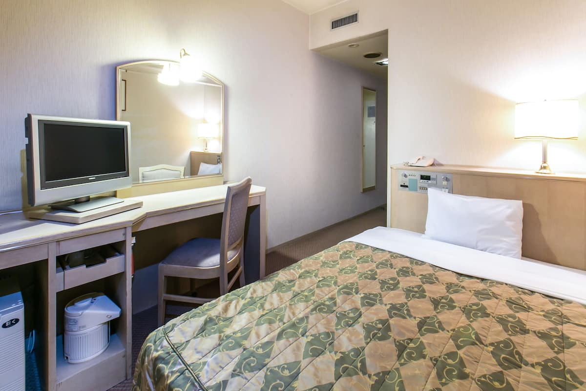 Hotellihuone, jossa on sänky, työpöytä tietokoneella, tuoli, yöpöytä ja lamppu.