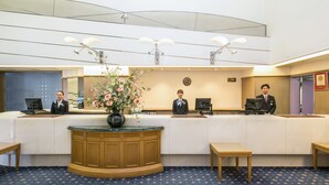 A hotel reception area with a reception desk, three staff members, a clock, and a vase with flowers.
