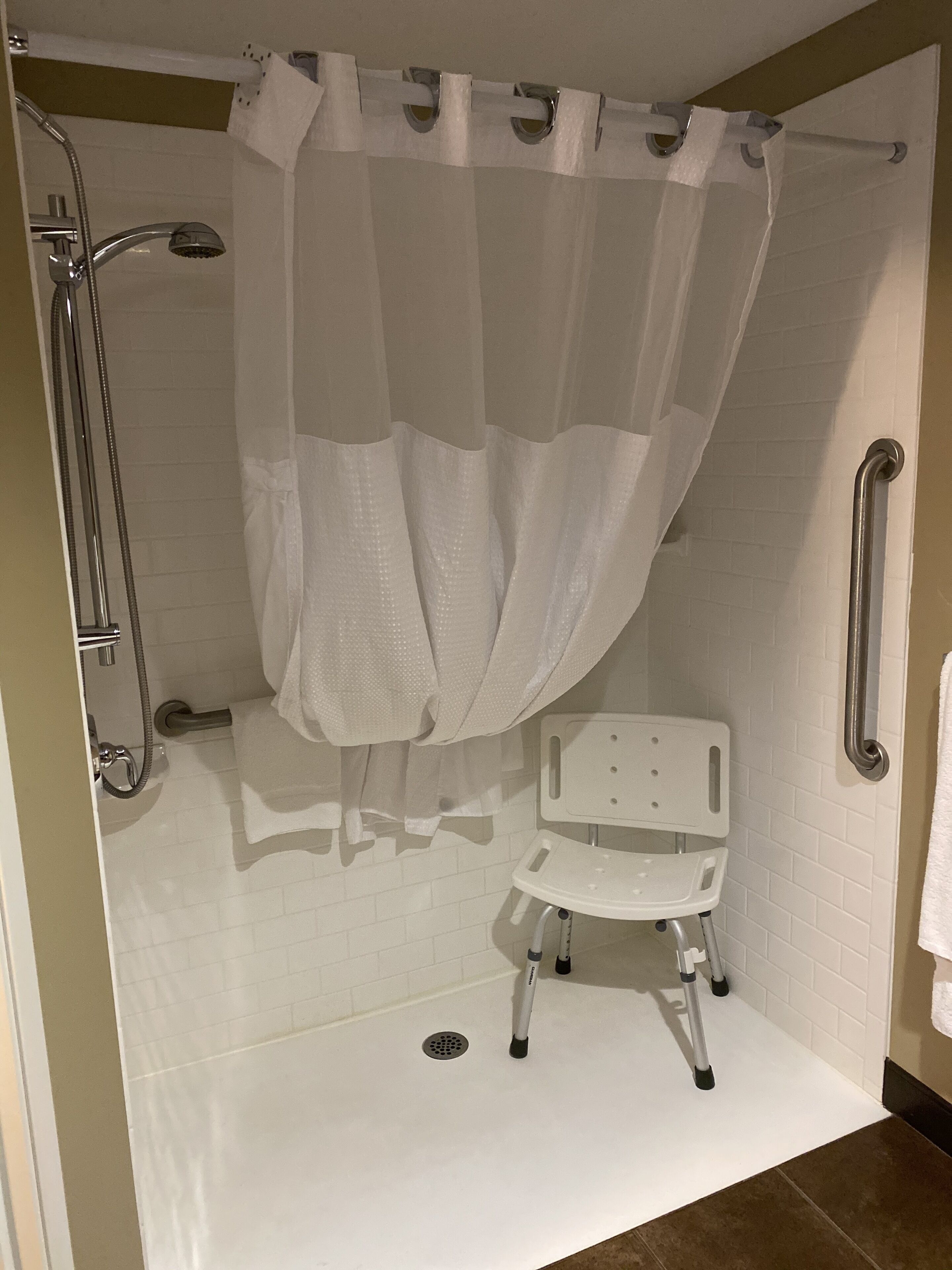 A shower area with a white shower curtain, grab bars, and a white mobility chair.