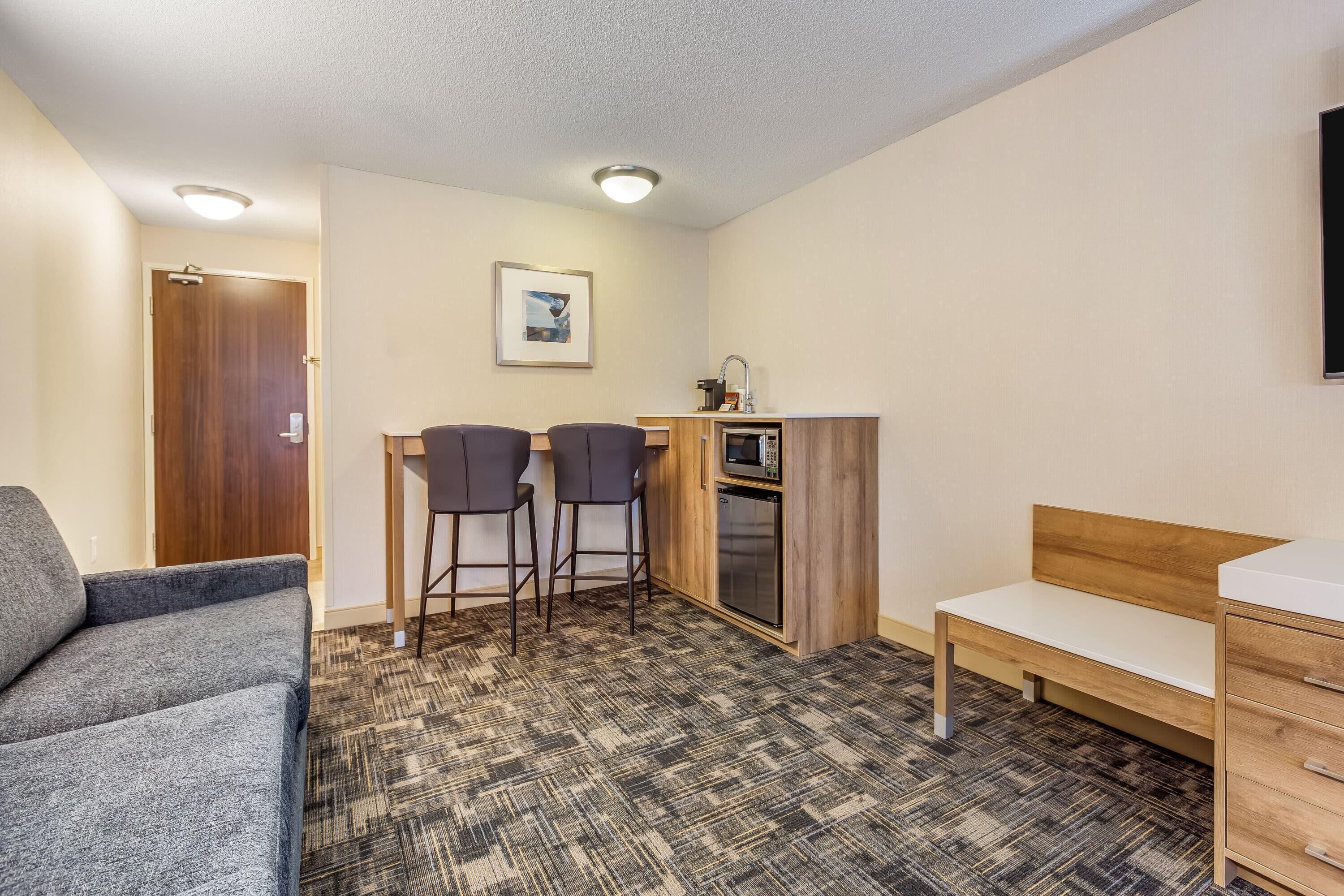 A compact room with a sofa, a small kitchenette, two bar stools, a wooden bench, and a single bed.
