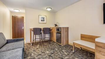 A compact room with a sofa, a small kitchenette, two bar stools, a wooden bench, and a single bed.