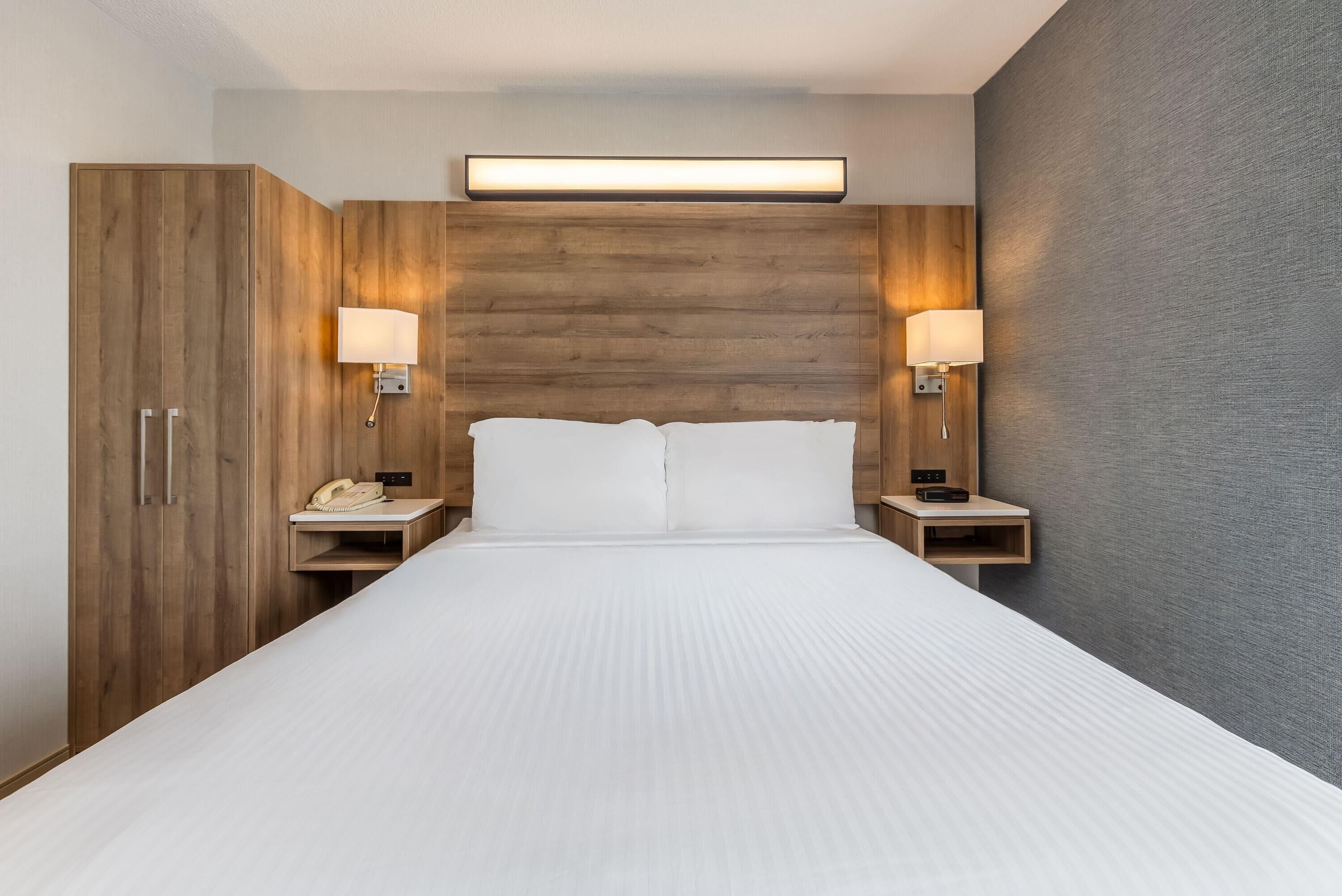 Quarto de hotel com uma cama grande, guarda-roupa de madeira, abajures e uma luminária na parede.