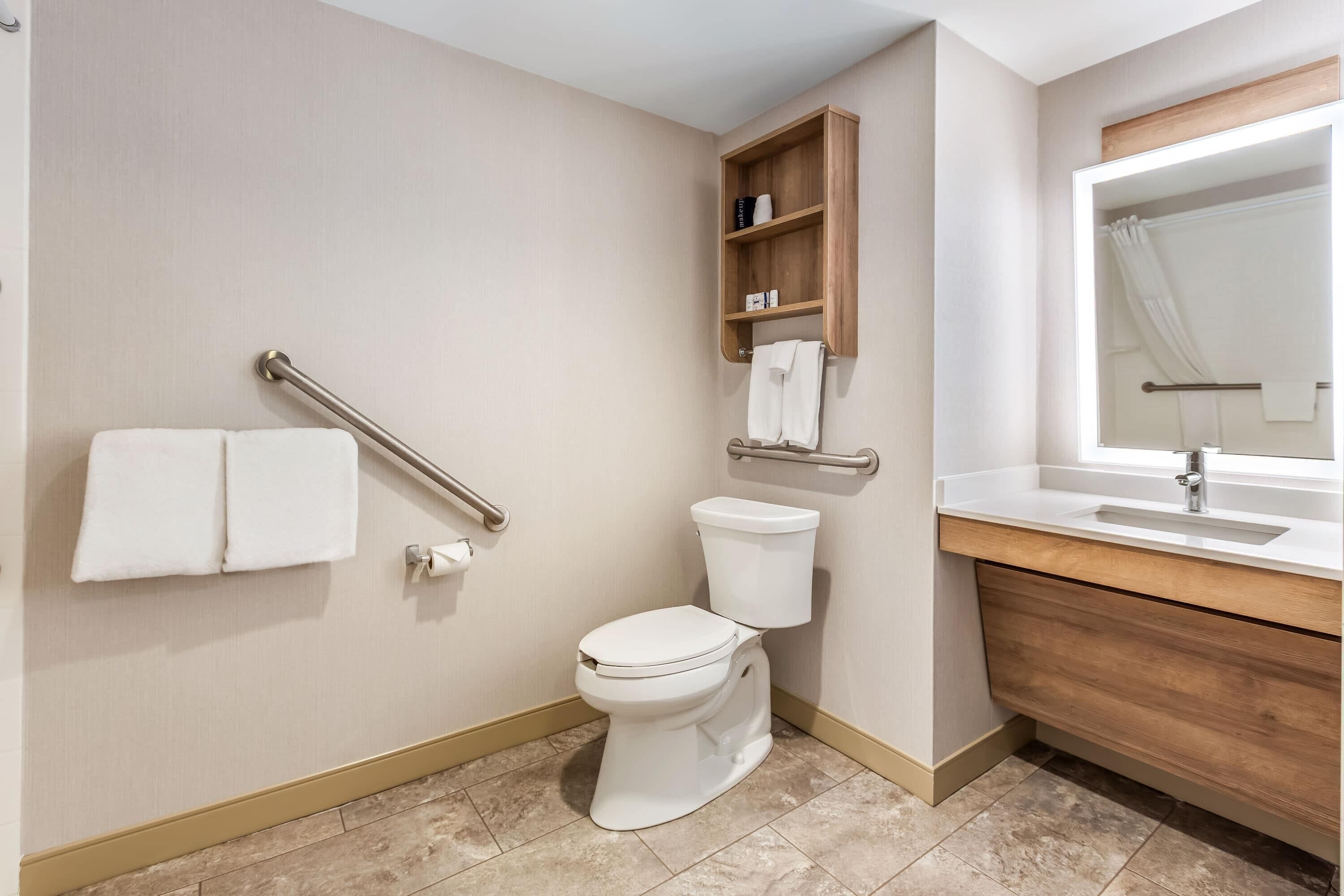 Ein Badezimmer mit Toilette, Handläufe, einem Ablagefach mit Handtüchern und einem Waschbecken mit Spiegel.