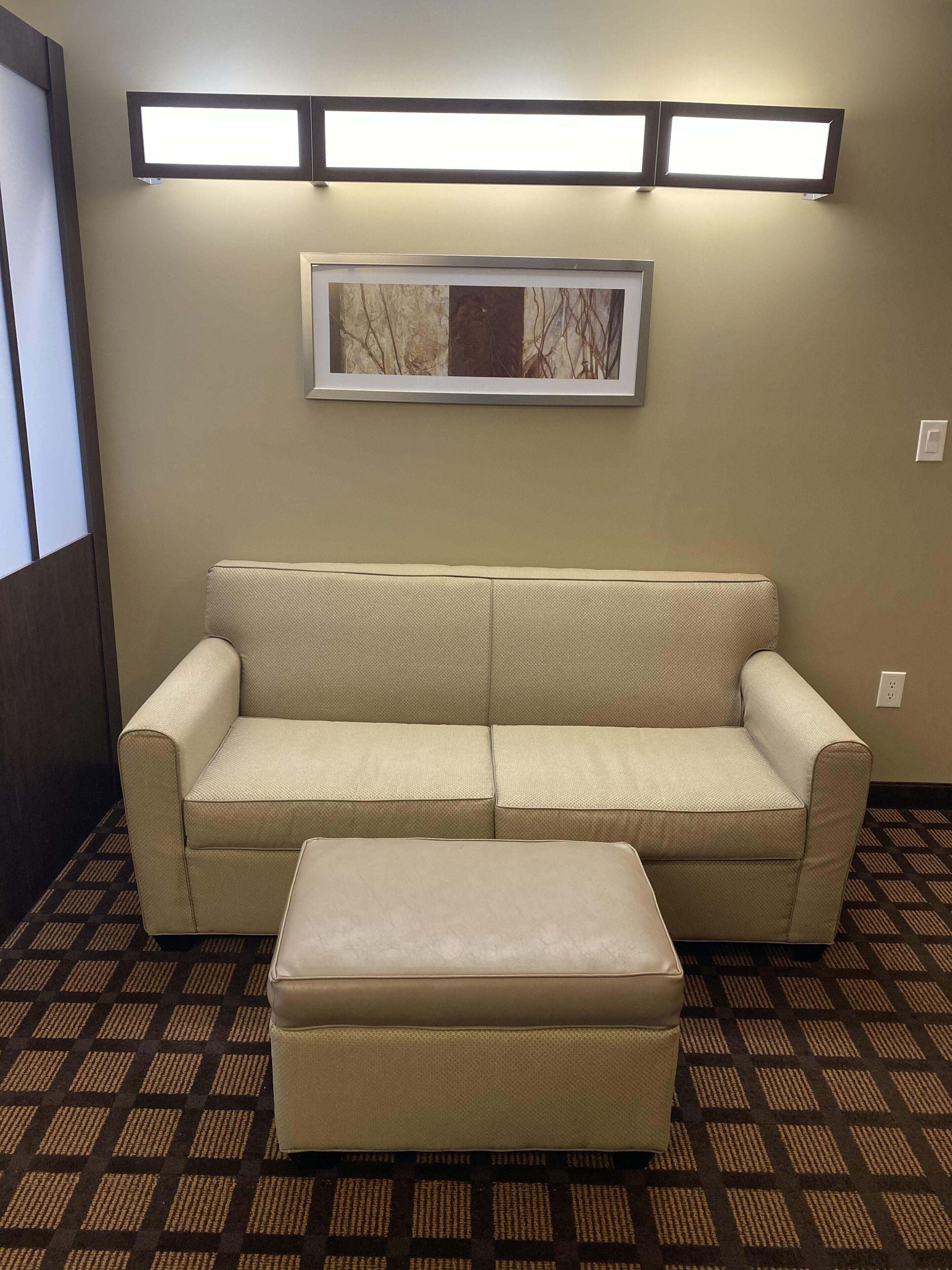 Ein beiges Sofa mit Hocker in einem Zimmer mit gemustertem Teppich, einem gerahmten Bild an der Wand und zwei Wandleuchten.