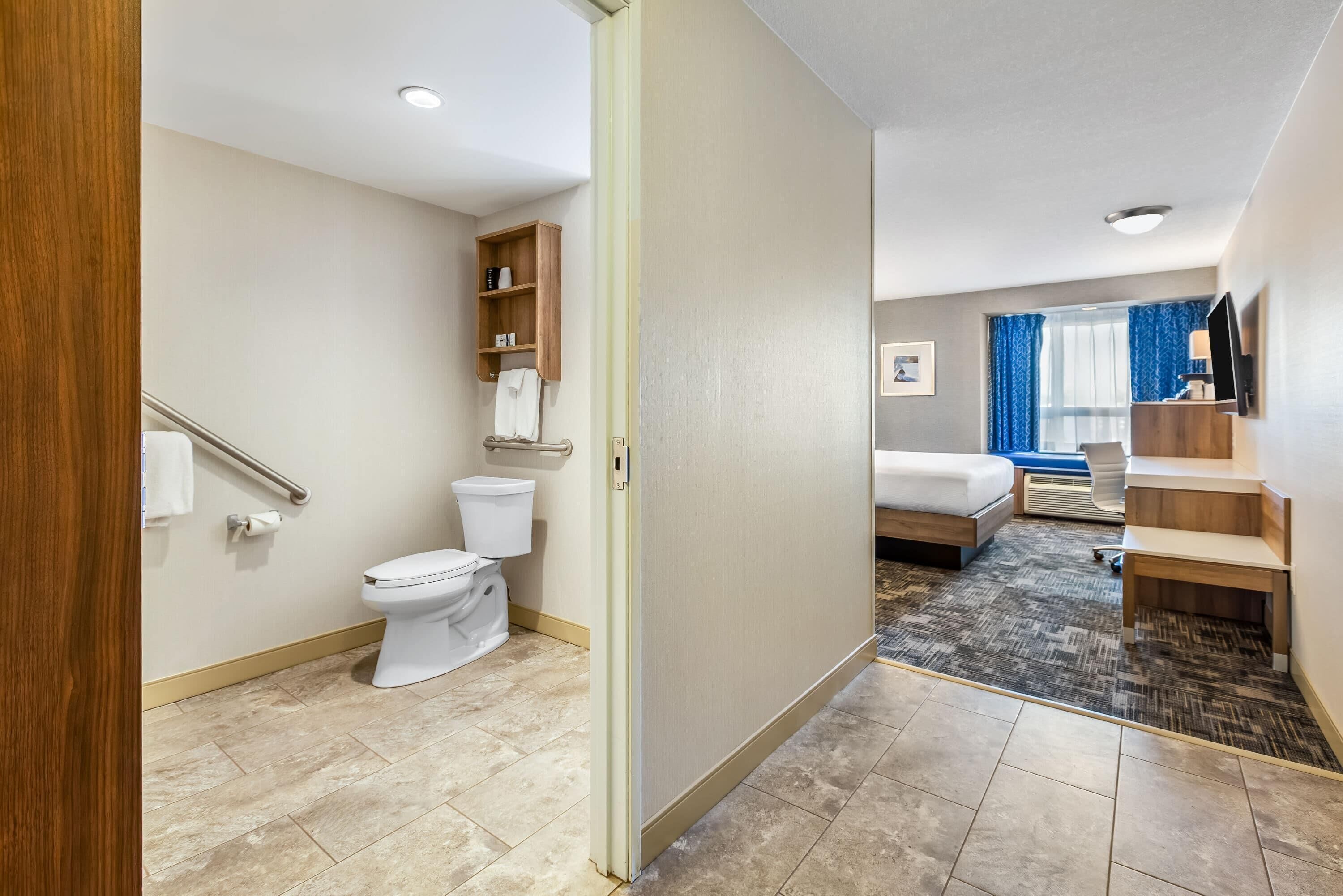 A hotel bathroom with a toilet, shelf, and a bed in the adjacent room.