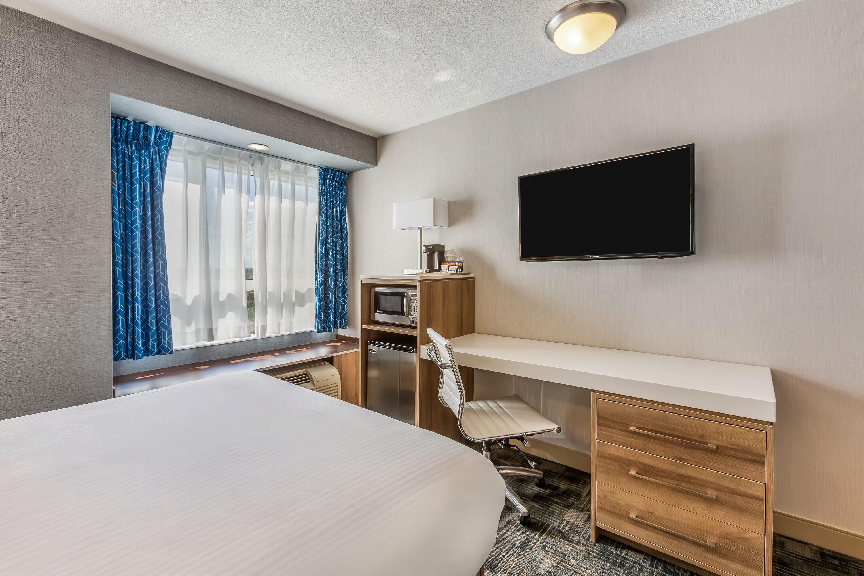 A hotel room with a bed, desk, chair, TV, microwave, and a window with curtains.