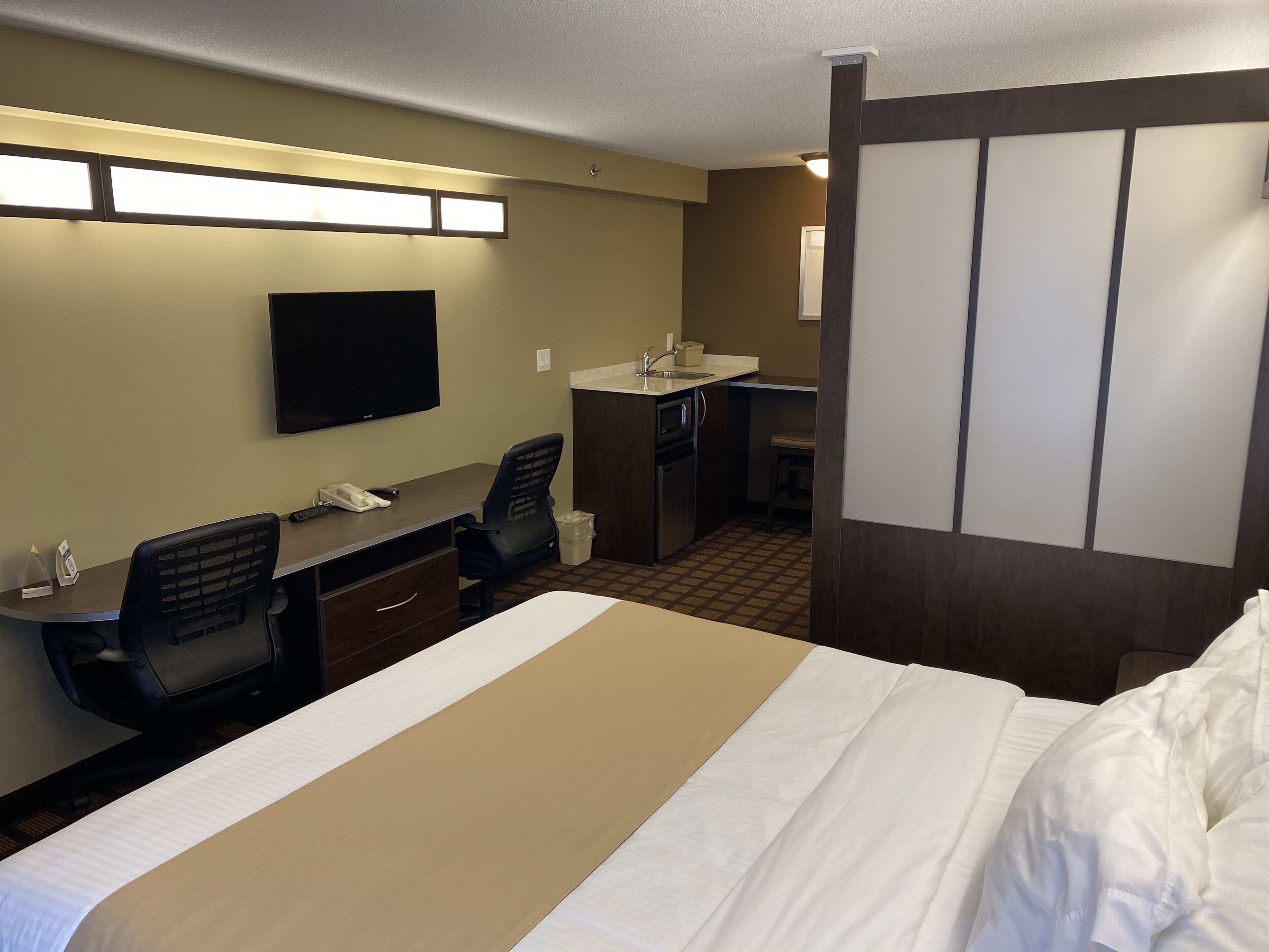A hotel room with a bed, desk, chair, television, and kitchenette.