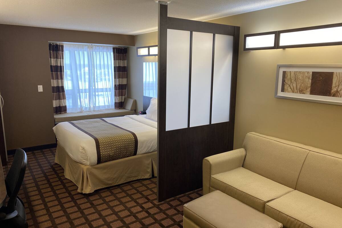 A hotel room with a bed, a sofa, a chair, a window with curtains, and a wall-mounted light fixture.