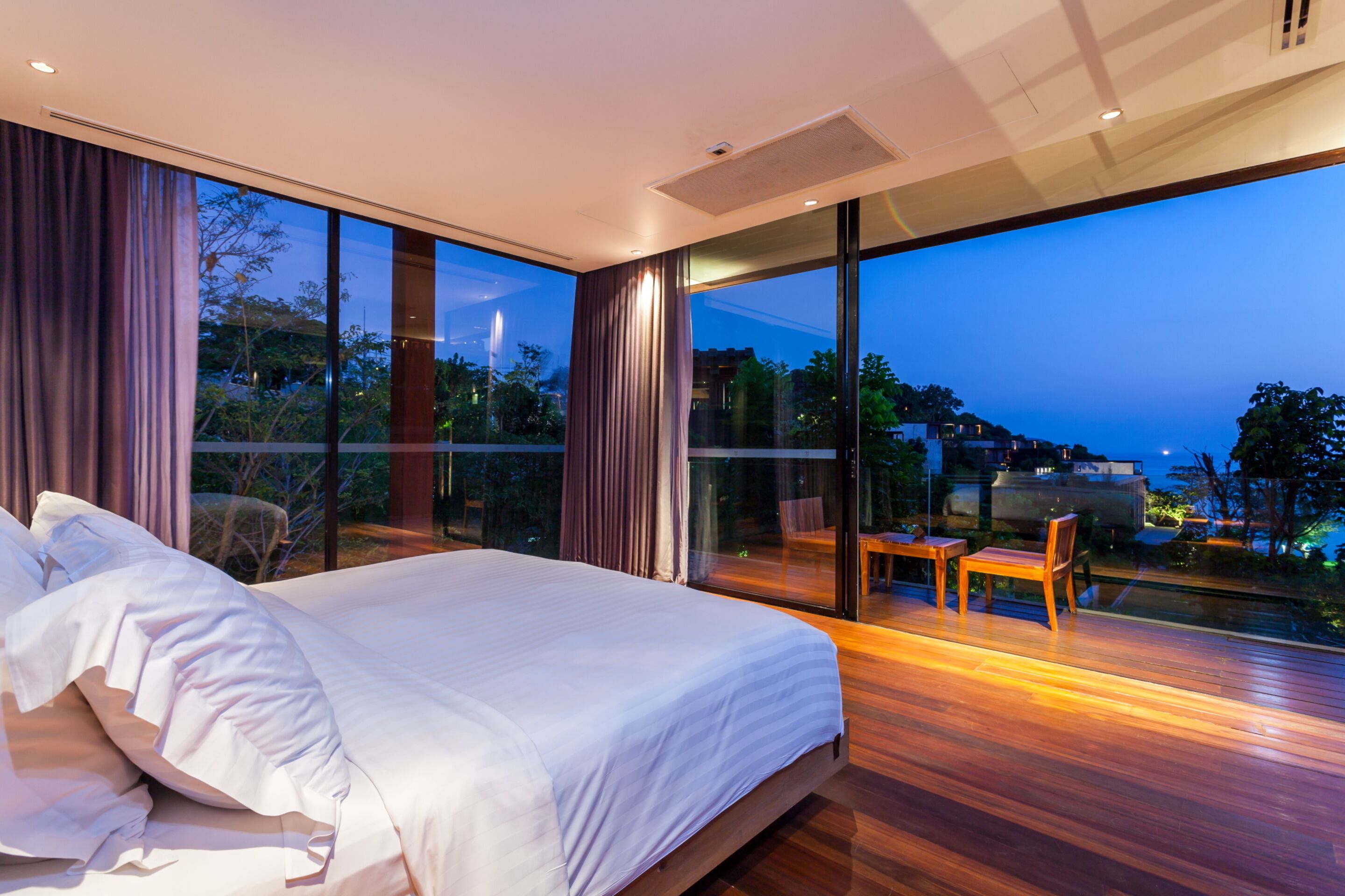 A modern bedroom with a large bed, a wooden floor, and a balcony with a view of the city at dusk.
