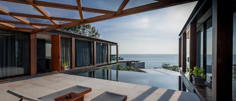 Un moderno espacio lounge al aire libre con piscina infinita de cristal, mobiliario de madera y vista al mar.