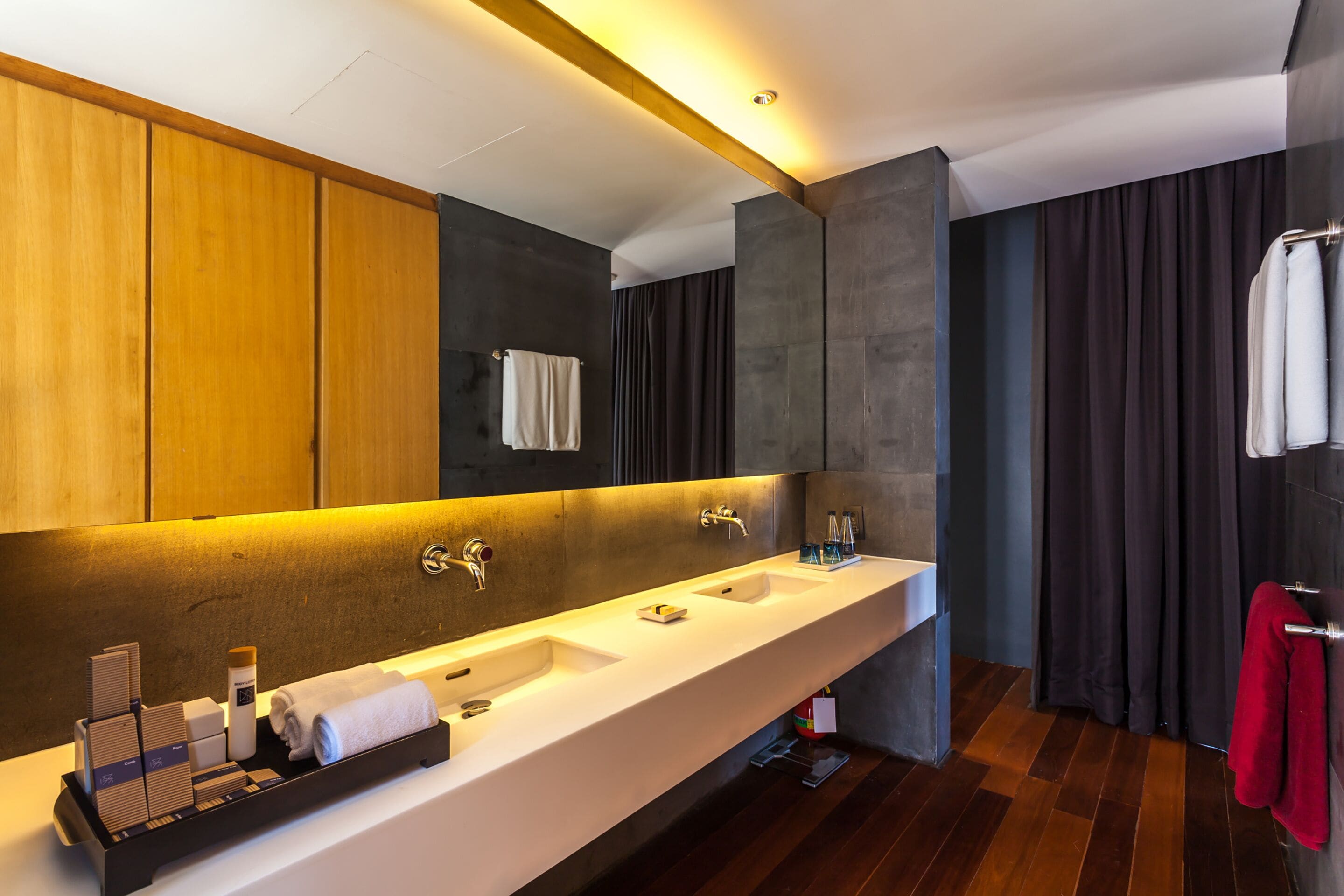 A modern bathroom with a long white sink, two faucets, and a large mirror. There are wooden cabinets and a dark curtain on one side.