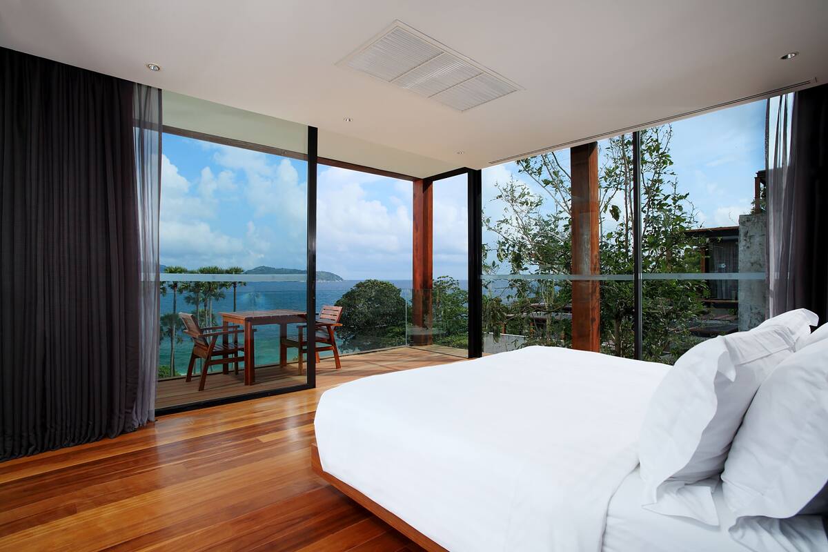 A modern bedroom with a large bed, a wooden floor, a balcony with a table and chairs, and a view of the sea.