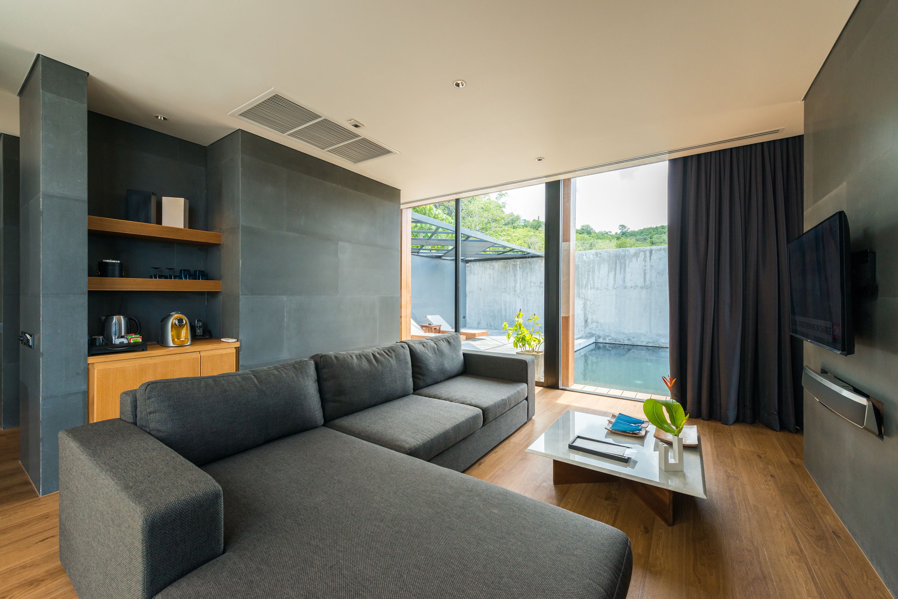 Un soggiorno moderno con un divano componibile, un tavolino da caffè e una TV. Si nota un mobile in legno contenente elettrodomestici da cucina e un'ampia finestra che si affaccia sul verde.