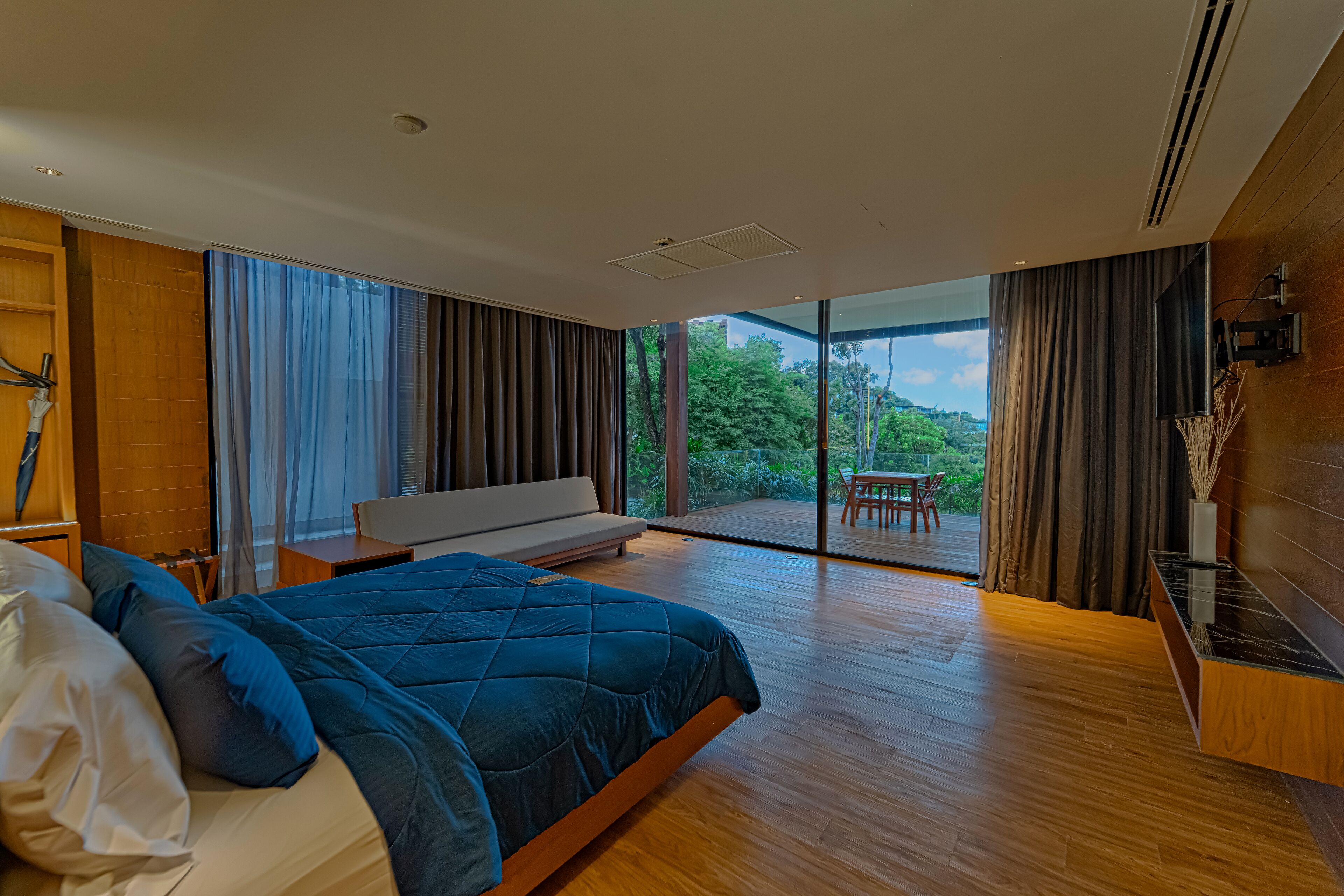 A modern bedroom with a large bed, a seating area, and a view of greenery through large windows.