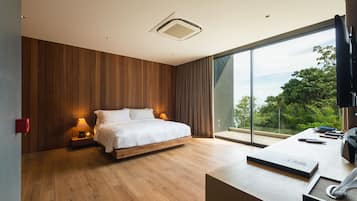 Un dormitorio moderno con una cama grande, paneles de madera, un escritorio con ordenador y vistas a la vegetación a través de amplias ventanas.