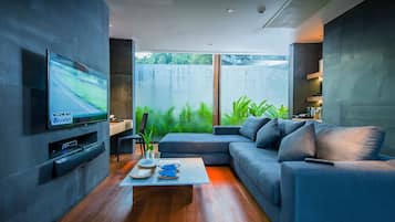 A modern living room with a flat-screen TV, a grey sofa, a coffee table, and a view of greenery outside.