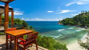 A wooden deck with a table and chairs overlooking a beach with clear blue waters and a rocky coastline.
