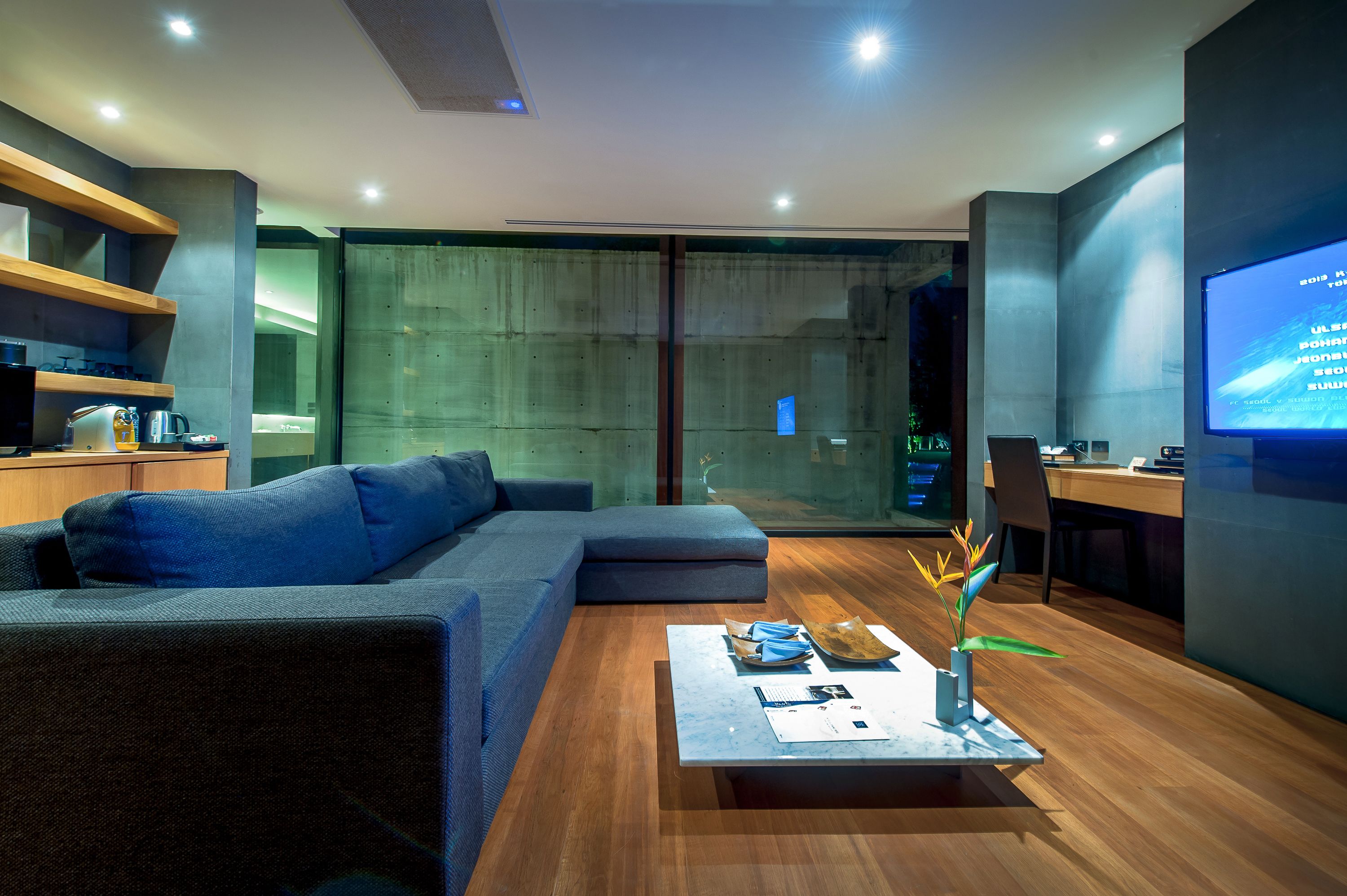 A modern living room with a sectional sofa, a coffee table with papers and a plant, a TV on the wall, and a glass door leading to a balcony.