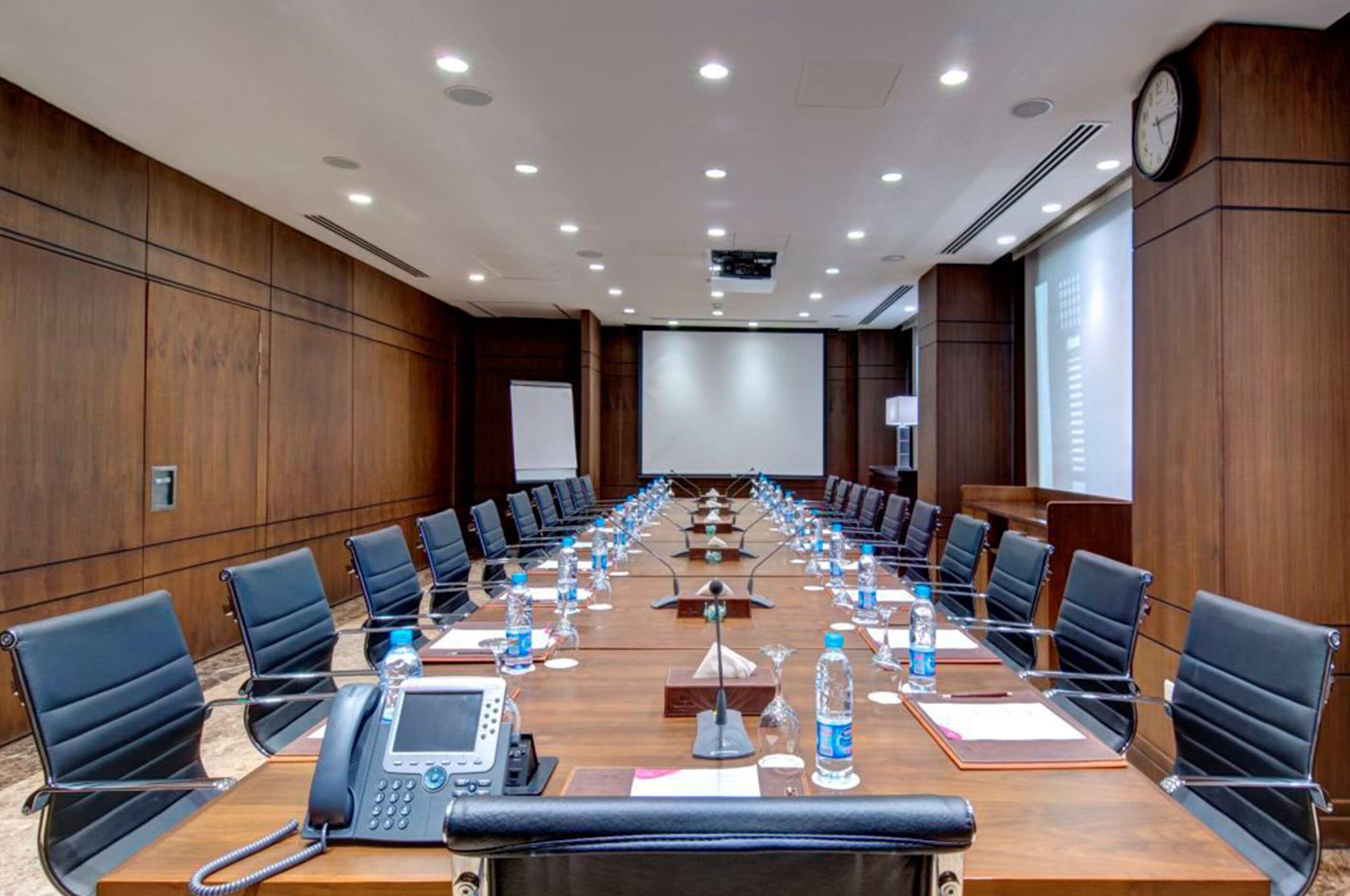 A conference room with a long table, chairs, a projector screen, and a clock on the wall.