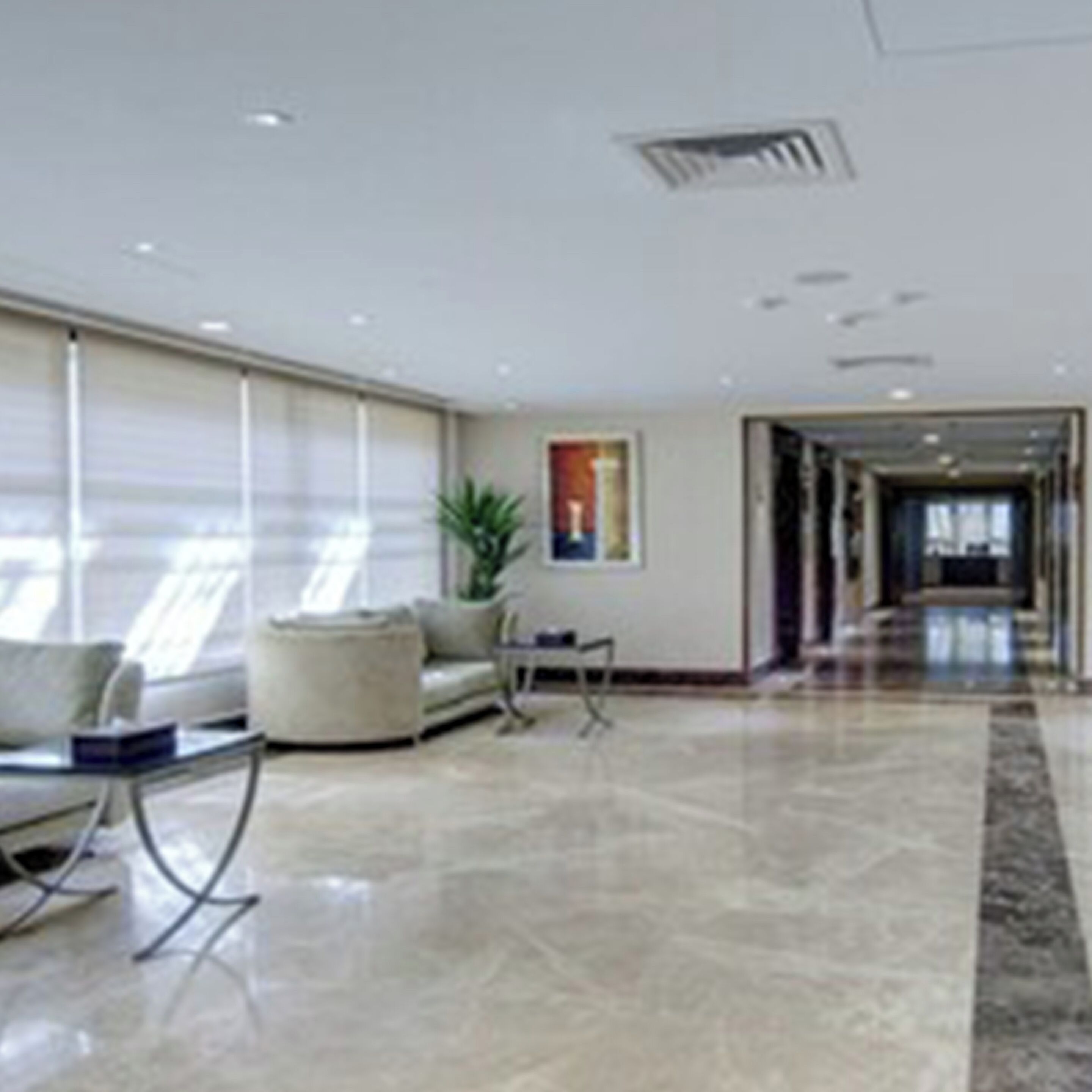 A spacious lobby with a modern design, featuring a seating area with sofas and a coffee table, a large window with blinds, and a hallway leading to other parts of the building.