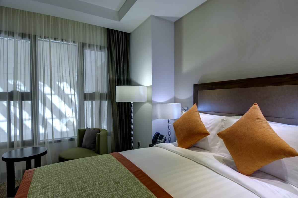 Ein Hotelzimmer mit Bett, zwei Kissen, einem Nachttisch, einer Lampe, einem Sessel und einem Fenster mit Vorhängen.