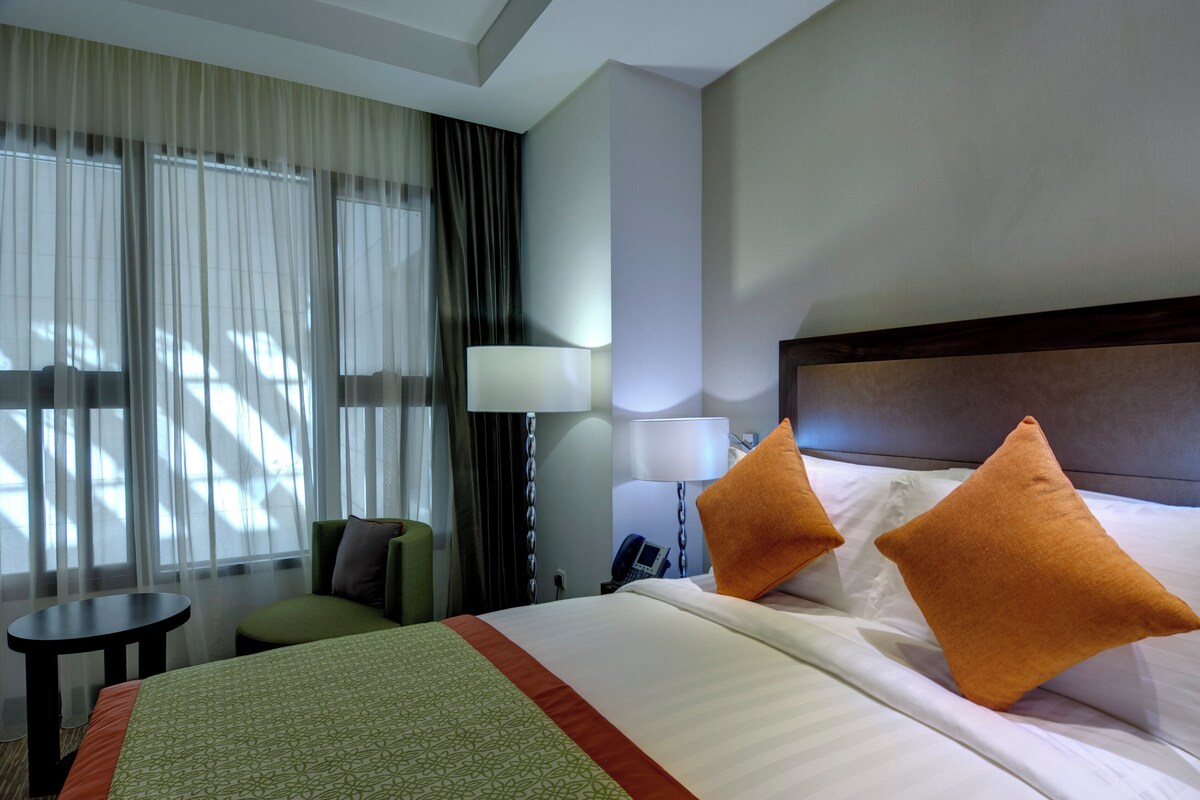 A hotel room with a bed, two pillows, a nightstand, a lamp, a chair, and a window with curtains.