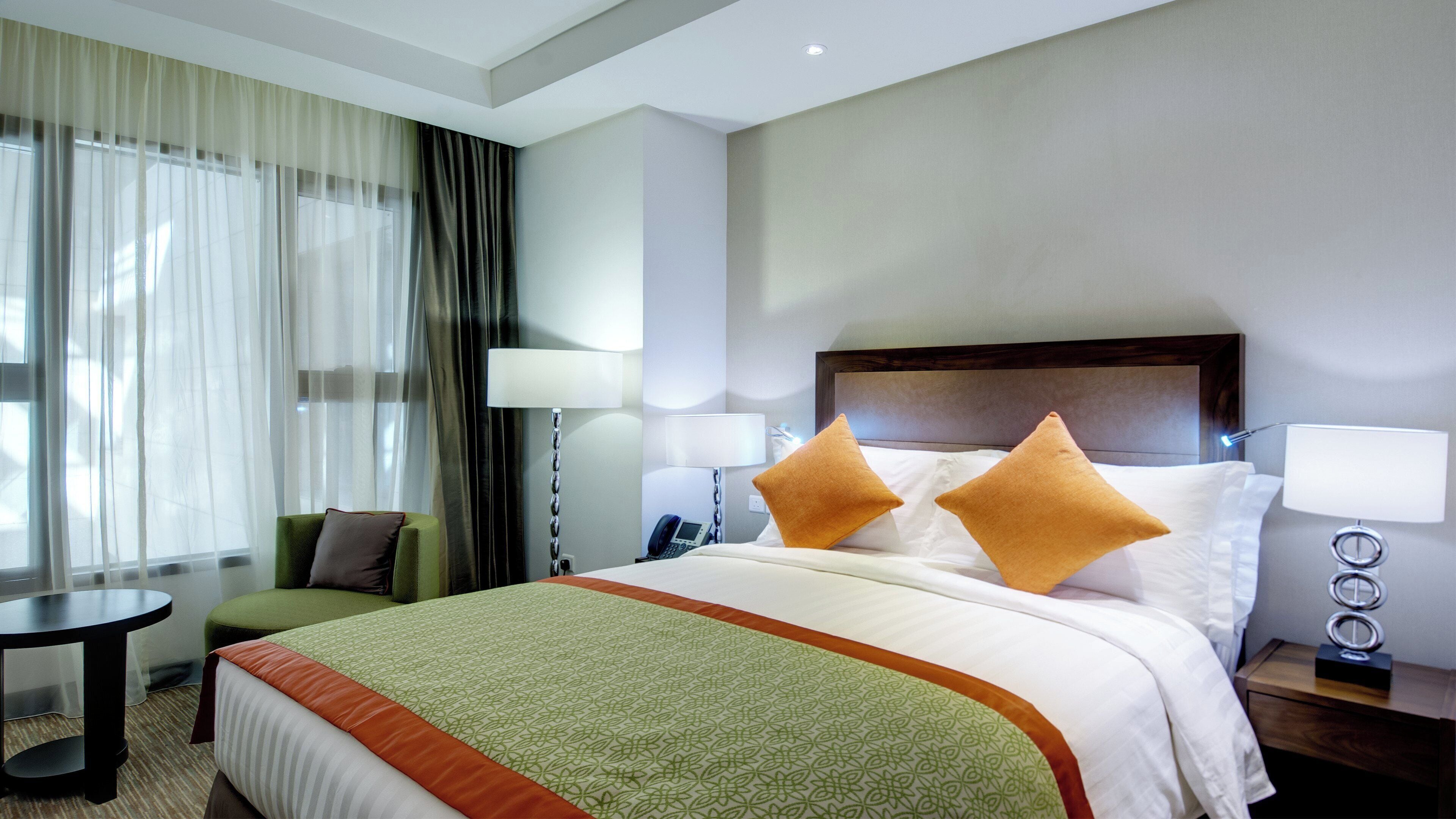 A hotel room with a bed, two pillows, a bedside table with a lamp, a chair, a desk, and a window with curtains.