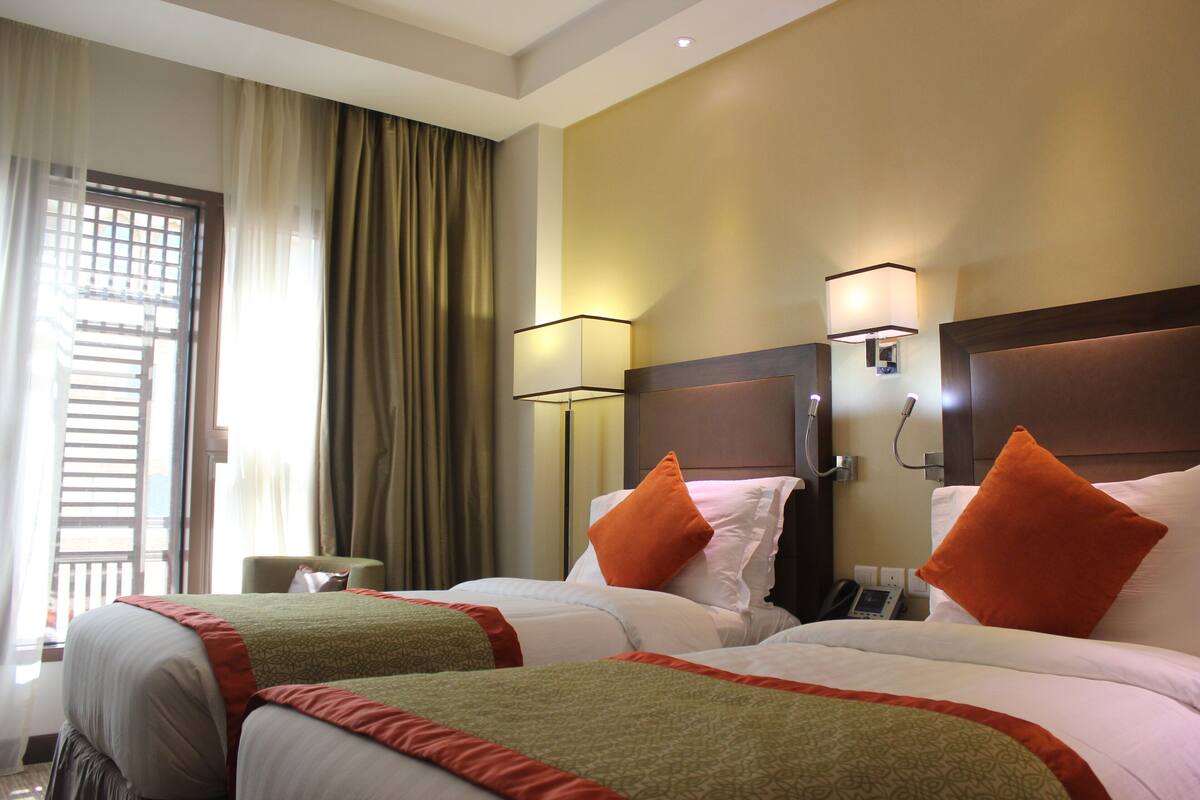 Ein Hotelzimmer mit zwei Betten, jeweils mit einer gemusterten Tagesdecke und orangefarbenen Kissen. An der Nachttischlampe befinden sich Wandlampen und ein Telefon.
