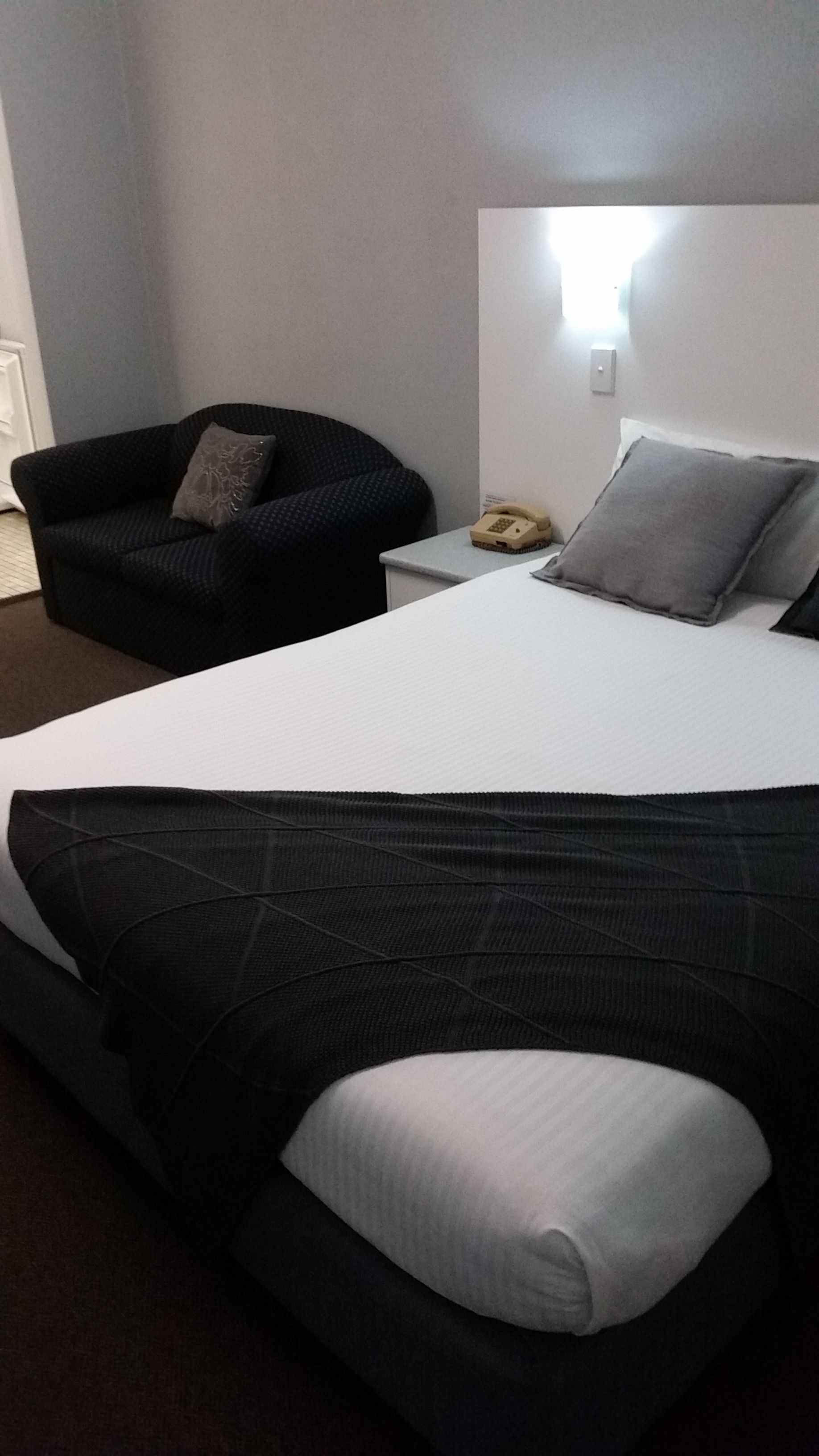 A hotel room with a large bed, a chair, a nightstand, and a wall-mounted light.